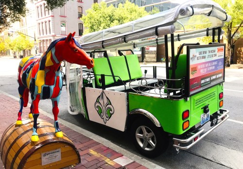 A colorful horse statue stands next to a green, open-air vehicle parked on a brick street.
