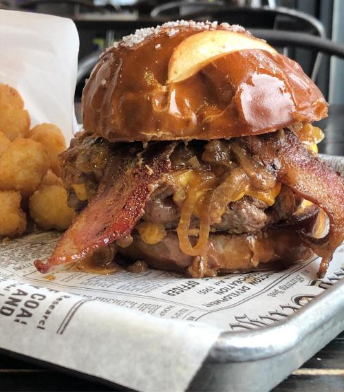 A close-up of a juicy burger with bacon, melted cheese, and caramelized onions on a pretzel bun, served with tater tots.