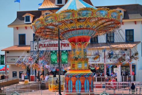 A colorful swing ride at a seaside amusement park, with people enjoying the attractions and a food stand nearby.