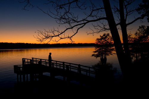 Silhouette of a person on a dock at sunset, with calm water and trees reflecting the vibrant orange and blue sky.