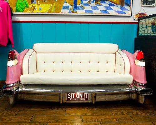 A vintage car-themed sofa with a white and pink design, featuring chrome accents and a "SIT ON IT" sign.