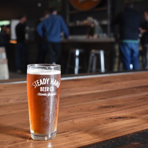 A glass of amber beer sits on a wooden bar, with blurred patrons in the background at a brewery.