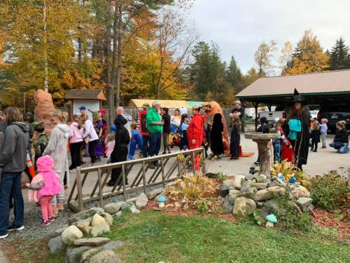 A crowd in costumes gathers outdoors among autumn trees, with decorations and a fountain in the foreground.