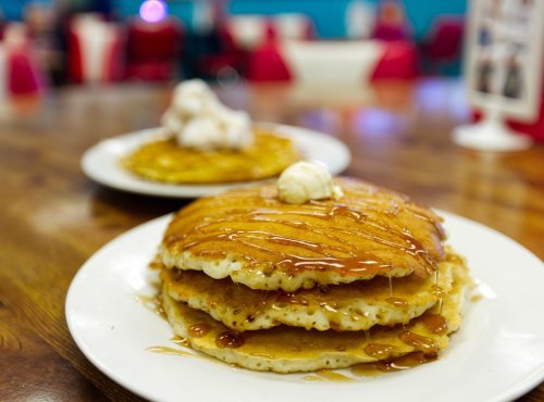 A stack of fluffy pancakes drizzled with syrup and topped with a dollop of butter, served on a white plate.