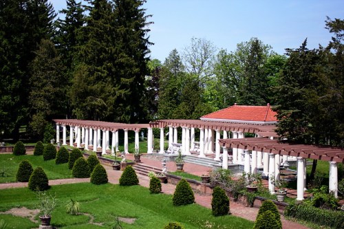 A serene garden with a colonnade, manicured hedges, and a red-roofed structure under a clear blue sky.