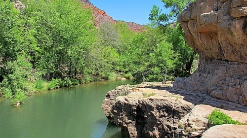 A serene river flows between lush greenery and rocky cliffs under a clear blue sky.