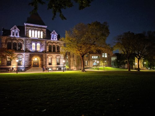 Historic building illuminated at night, surrounded by trees and a grassy area, with people walking nearby.