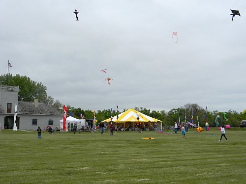 A grassy field with people flying kites near a large yellow tent on a cloudy day.