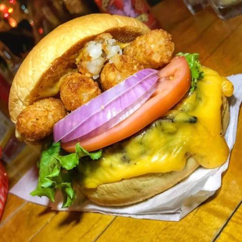 A delicious burger with melted cheese, lettuce, tomato, onion, and crispy tater tots on a toasted bun.