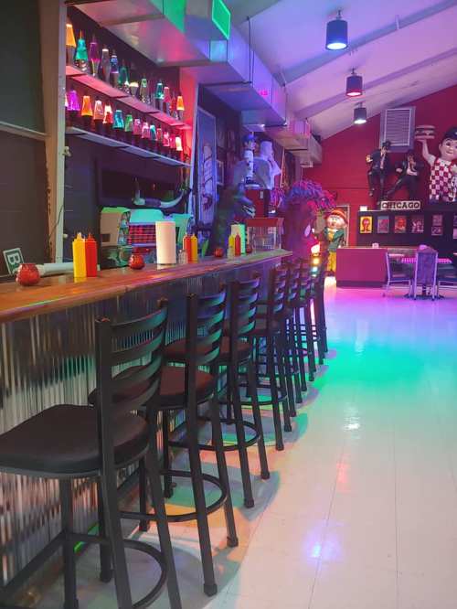 A colorful bar with empty black chairs, vibrant decor, and playful lighting in a lively setting.