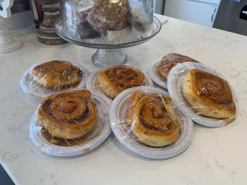 A group of cinnamon rolls wrapped in plastic on white plates, with a glass cake stand of baked goods in the background.