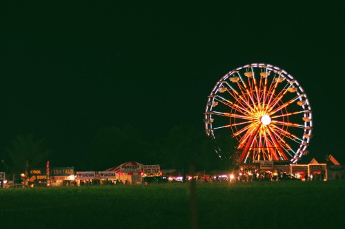 A brightly lit ferris wheel at night, surrounded by food stalls and a grassy field.