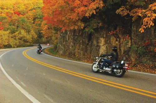 Motorcyclists ride along a winding road surrounded by vibrant autumn foliage.