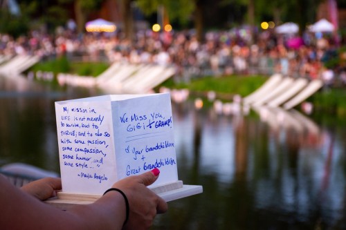 A person holds a handwritten tribute near a crowd by a lake, expressing love and remembrance for a loved one.