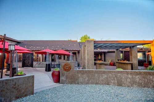 Outdoor dining area with red umbrellas, modern architecture, and a gravel landscape under a clear blue sky.