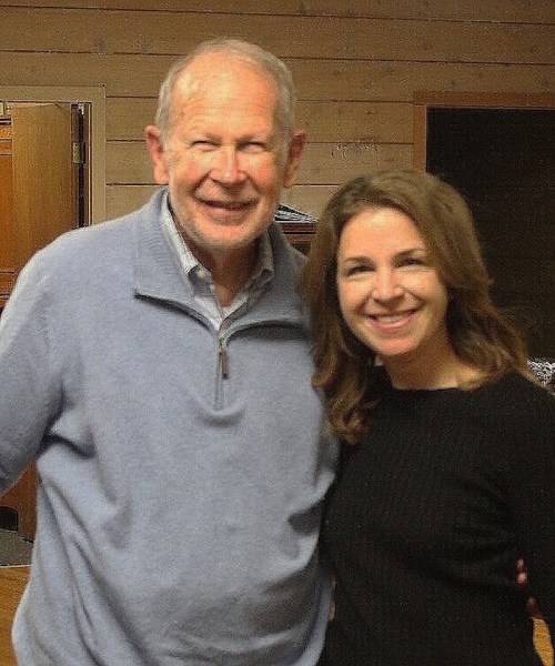 A smiling man and woman pose together indoors, both wearing casual clothing.