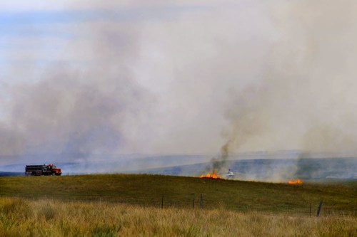 Firefighters respond to a grass fire on a hillside, with smoke billowing and flames visible in the foreground.
