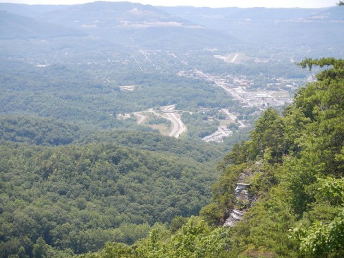 A panoramic view of a valley with a winding road, surrounded by lush green mountains and a small town in the distance.