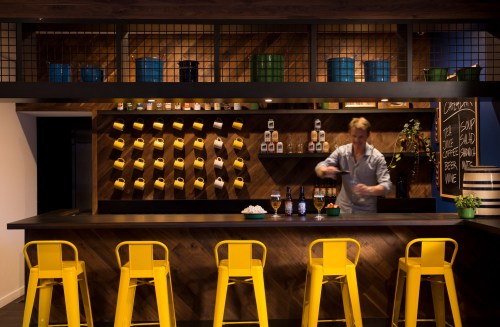 A modern bar with yellow stools, a bartender mixing drinks, and shelves displaying various bottles and containers.
