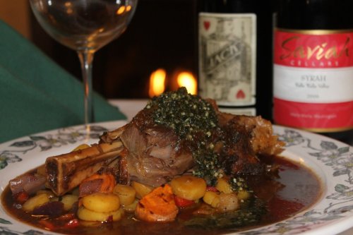 A plate of lamb shank with vegetables, garnished with sauce, alongside wine bottles and a glass. Warm ambiance in the background.