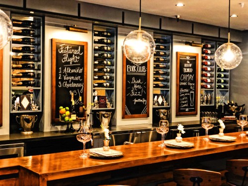 A stylish bar with wooden counter, wine bottles on display, and chalkboard menus featuring drinks and specials.