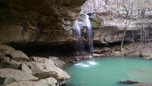 A serene waterfall cascades into a turquoise pool, surrounded by rocky cliffs and bare trees in a tranquil forest setting.