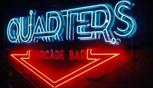 Neon sign for "Quarters Arcade Bar" with vibrant blue and red lighting against a dark background.