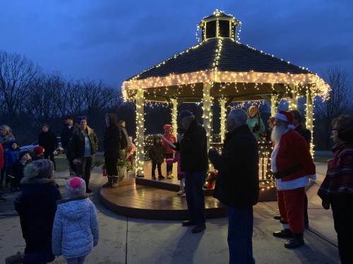 A festive gathering around a lit gazebo with people, including Santa, celebrating in a park during twilight.