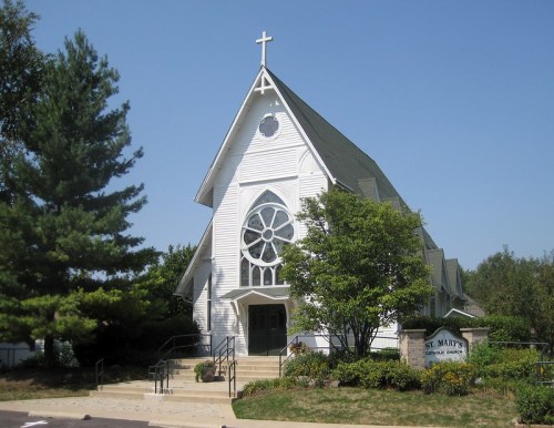 A white church with a cross on top, featuring a large circular window and surrounded by trees and greenery.
