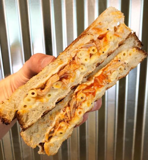 A hand holding a grilled cheese sandwich filled with macaroni and cheese and shredded chicken, against a metallic background.
