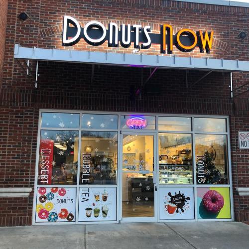 Exterior of "Donuts Now" shop with a bright sign, open door, and displays for donuts and bubble tea.