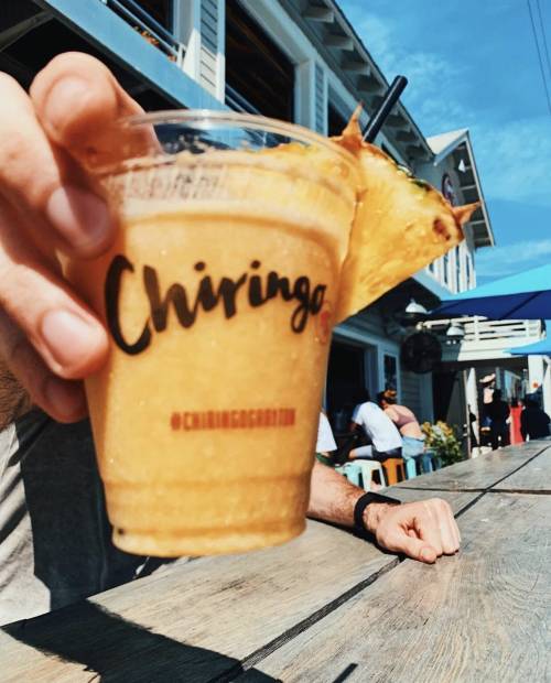 A person holds a frozen drink with a pineapple slice, featuring the word "Chiringo" on the cup, against a sunny backdrop.