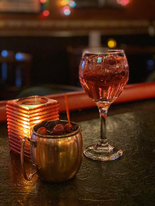 A copper mug filled with cherries and a glass of red drink sit on a bar, illuminated by soft candlelight.