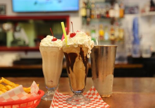 Two tall milkshakes topped with whipped cream and cherries, alongside a metal shaker, with fries in a basket in the foreground.