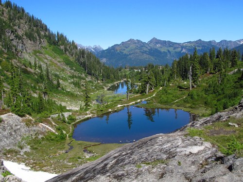 A serene mountain landscape featuring a calm pond surrounded by lush greenery and distant peaks under a clear blue sky.