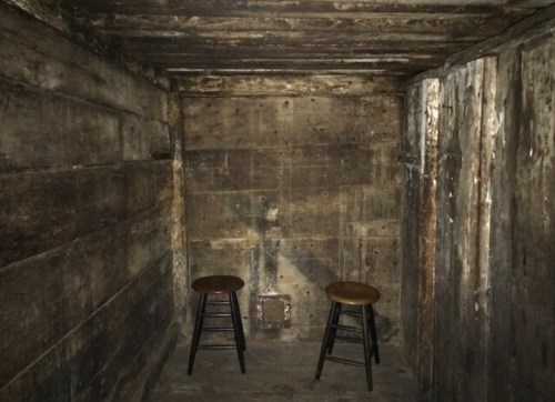A dimly lit, empty room with rough wooden walls and two wooden stools positioned on the floor.