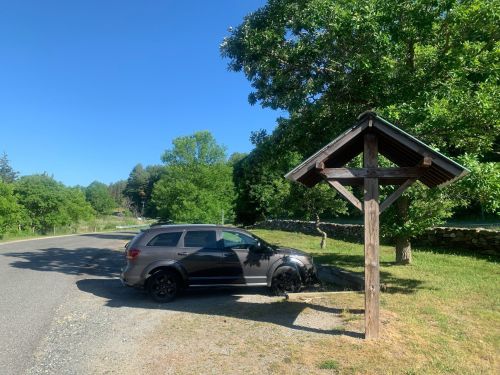 A parked car beside a wooden signpost under a tree, with a clear blue sky and green landscape in the background.