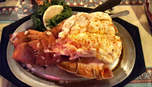A plated lobster tail topped with butter and spices, garnished with lemon and lettuce on a metal serving dish.