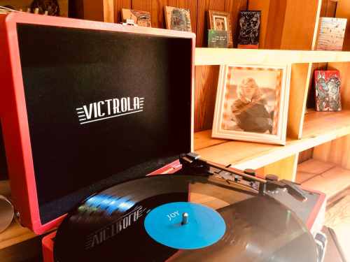 A red Victrola record player with a vinyl record labeled "Joy," set against a wooden shelf with framed photos.