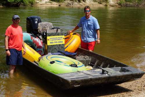 Two men stand on a boat by the river, surrounded by colorful inflatable tubes and a sign for Louisiana River Adventures.