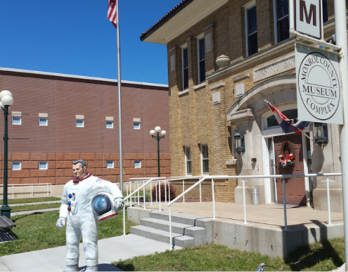 A life-sized astronaut statue stands outside the Monroeville Museum Complex, with a flag and building in the background.