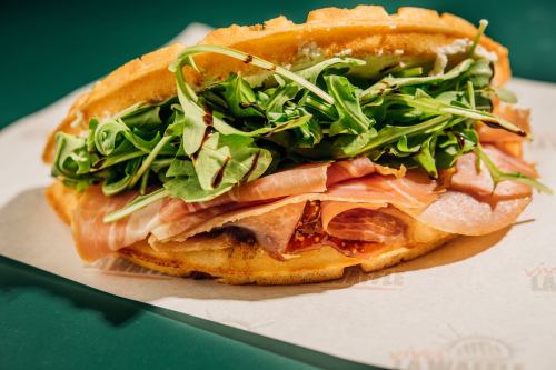 A close-up of a sandwich with layers of greens, ham, and other meats, served on a waffle-like bread.