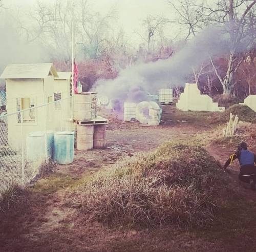 A paintball field with smoke rising, featuring structures and a player crouching behind a mound of grass.