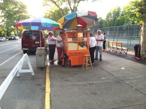 A popcorn stand with colorful umbrellas, surrounded by people, parked beside a road with benches and trees in the background.