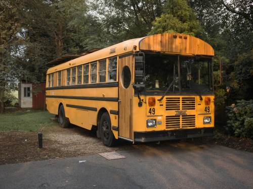 A vintage yellow school bus parked on a gravel driveway surrounded by trees and a house in the background.