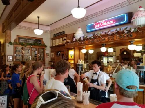 A busy ice cream shop with a line of customers and a server preparing orders at the counter. Bright, inviting atmosphere.