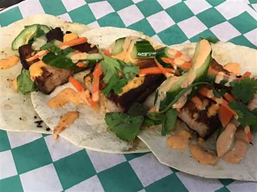 Three tacos filled with grilled meat, topped with vegetables and drizzled with sauce, served on a green checkered background.