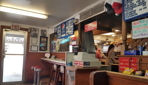Interior of a diner with a counter, stools, and a menu board, featuring a vintage atmosphere and staff behind the counter.