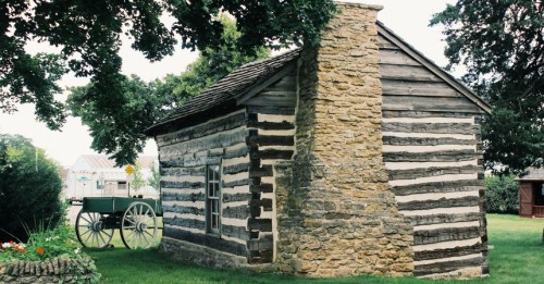 A rustic log cabin with a stone chimney, surrounded by greenery and a vintage wagon nearby.