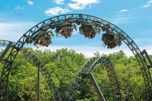 A roller coaster with riders upside down, set against a backdrop of green trees and a blue sky.